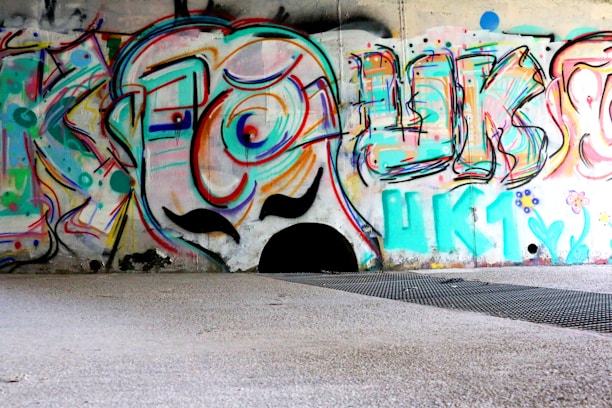 Colorful graffiti art covering a wall in an urban setting. The artwork features abstract shapes, swirling lines, and bold letters with a cartoonish face in the center. The vibrant colors include shades of blue, pink, orange, and green. The ground in front of the wall is a mixture of concrete and metal grating.