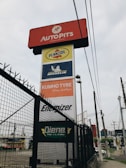 A tall signage tower displays multiple brand logos and names. Brands like Autopits, Pennzoil, Michelin, Kumho Tyre, Energizer, and others are shown on individual panels stacked vertically. The structure is surrounded by a black wire fence, with power lines visible in the background, set against a cloudy sky.