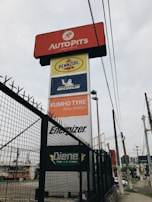 A tall signage tower displays multiple brand logos and names. Brands like Autopits, Pennzoil, Michelin, Kumho Tyre, Energizer, and others are shown on individual panels stacked vertically. The structure is surrounded by a black wire fence, with power lines visible in the background, set against a cloudy sky.