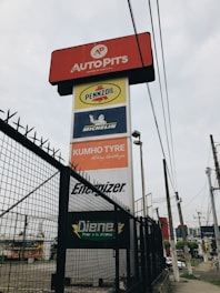 A tall signage tower displays multiple brand logos and names. Brands like Autopits, Pennzoil, Michelin, Kumho Tyre, Energizer, and others are shown on individual panels stacked vertically. The structure is surrounded by a black wire fence, with power lines visible in the background, set against a cloudy sky.