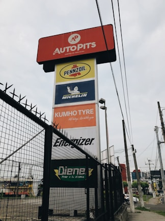 A tall signage tower displays multiple brand logos and names. Brands like Autopits, Pennzoil, Michelin, Kumho Tyre, Energizer, and others are shown on individual panels stacked vertically. The structure is surrounded by a black wire fence, with power lines visible in the background, set against a cloudy sky.