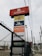 A tall signage tower displays multiple brand logos and names. Brands like Autopits, Pennzoil, Michelin, Kumho Tyre, Energizer, and others are shown on individual panels stacked vertically. The structure is surrounded by a black wire fence, with power lines visible in the background, set against a cloudy sky.