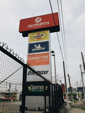 A tall signage tower displays multiple brand logos and names. Brands like Autopits, Pennzoil, Michelin, Kumho Tyre, Energizer, and others are shown on individual panels stacked vertically. The structure is surrounded by a black wire fence, with power lines visible in the background, set against a cloudy sky.
