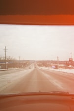 A road extending into the distance, flanked by buildings and utility poles, viewed from a car's windshield with a reddish tint at the top. The road and surrounding area appear to be lightly covered in snow.