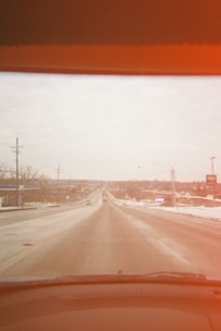 A road extending into the distance, flanked by buildings and utility poles, viewed from a car's windshield with a reddish tint at the top. The road and surrounding area appear to be lightly covered in snow.