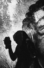 A shadow of a person holding an object is cast against a textured and possibly cracked surface. The contrast between the dark silhouette and the lighter background creates a striking visual effect.