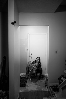 A woman is sitting on the floor in a hallway, embracing a large dog in front of a closed door. The setting is indoors with a simple and narrow layout, and household items are visible around the edges.