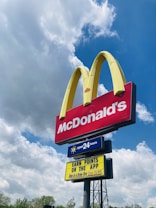 A McDonald's sign stands prominently against a partly cloudy blue sky. The large yellow arches and red background are easily recognizable. Beneath it, smaller signs advertise the restaurant being open 24 hours and a promotion to earn points through an app.