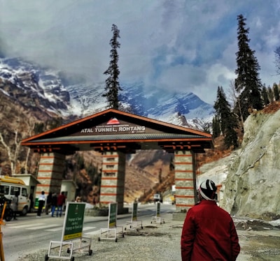 An entrance to the Atal Tunnel in Rohtang is visible, with signage marking the location. Snow-capped mountains and tall pine trees provide a scenic backdrop, while a person in a red jacket stands in the foreground. Several people are gathered near the entrance, and vehicles are parked on the side.