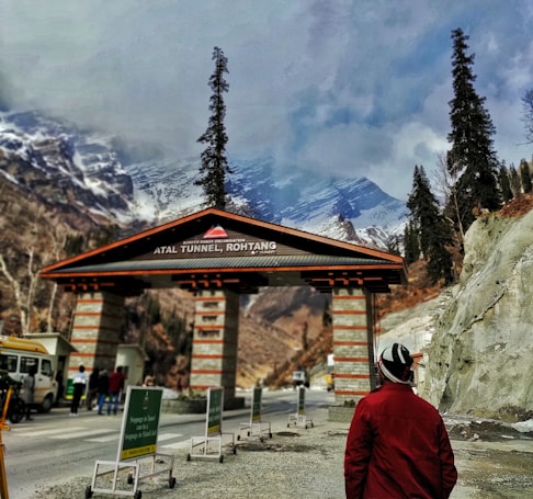 An entrance to the Atal Tunnel in Rohtang is visible, with signage marking the location. Snow-capped mountains and tall pine trees provide a scenic backdrop, while a person in a red jacket stands in the foreground. Several people are gathered near the entrance, and vehicles are parked on the side.