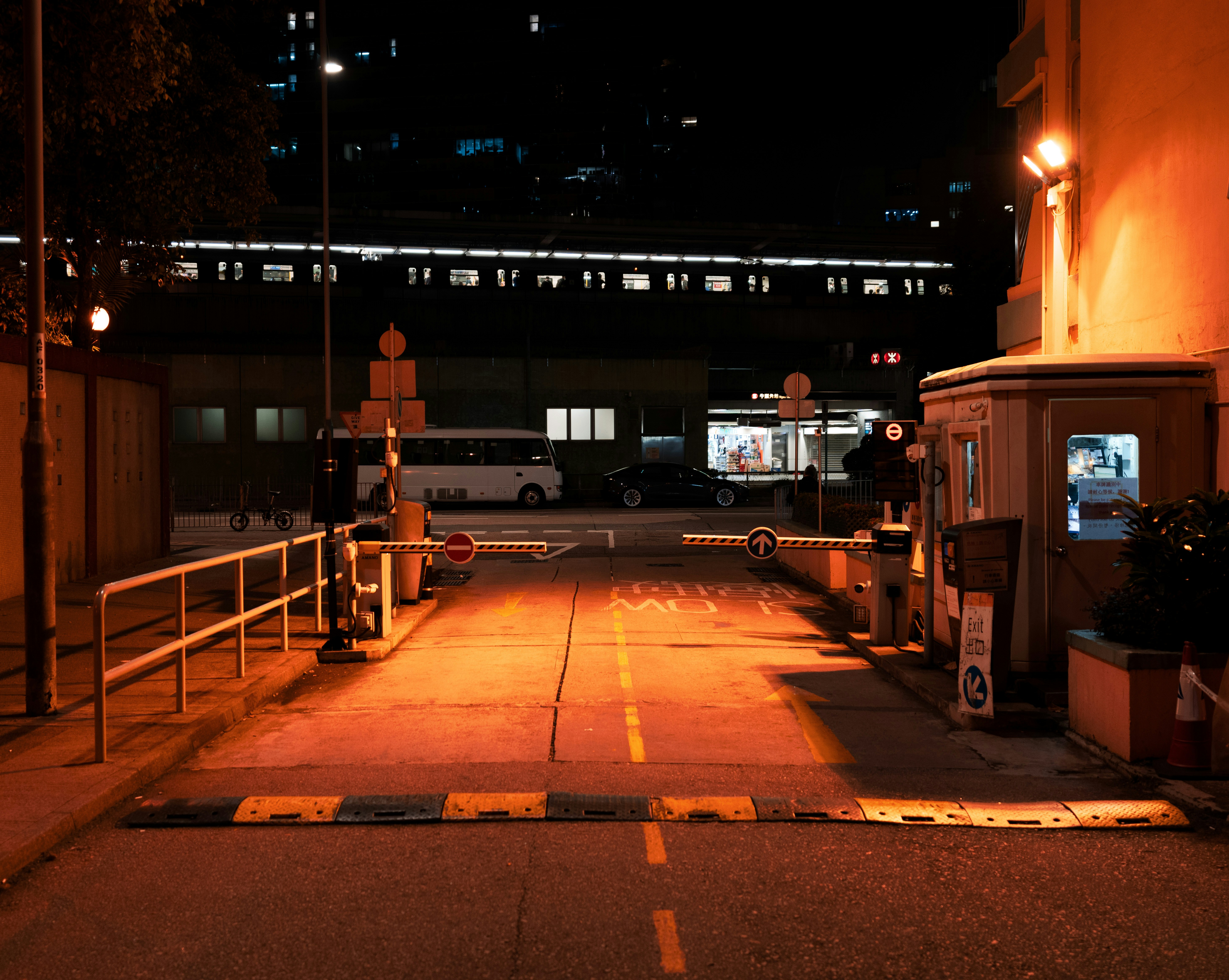Gated society entrance at night, dark ambient lighting, security booth with dim warm glow, deep shadows on boundary walls