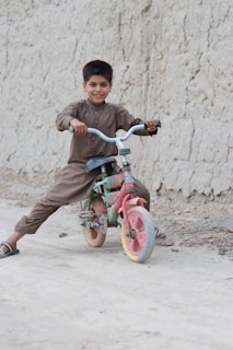 A young boy is smiling while sitting on a colorful bicycle. He is wearing a traditional brown outfit and sandals, with the bicycle positioned on a dirt path beside a textured wall.