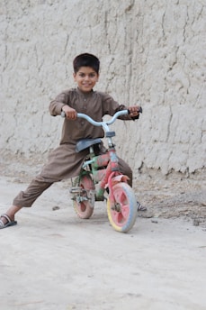 A young boy is smiling while sitting on a colorful bicycle. He is wearing a traditional brown outfit and sandals, with the bicycle positioned on a dirt path beside a textured wall.