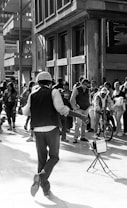 A street performer is energetically dancing in front of a crowd on a busy city street. The performer is wearing a vest and a hat. People around are watching and some are taking photos. A child on a bicycle is visible among the spectators.