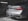 A luxury car in a dimly-lit garage is being washed, with soap suds covering the back of the vehicle. The car is silver and features distinctive red taillights and dual exhaust pipes. A license plate is visible, and cleaning equipment can be seen in the garage background.