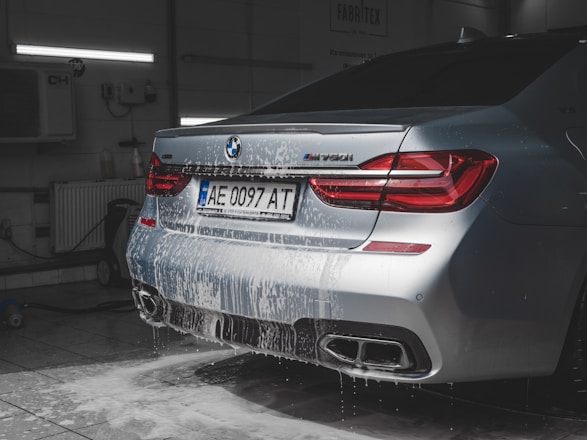 A luxury car in a dimly-lit garage is being washed, with soap suds covering the back of the vehicle. The car is silver and features distinctive red taillights and dual exhaust pipes. A license plate is visible, and cleaning equipment can be seen in the garage background.