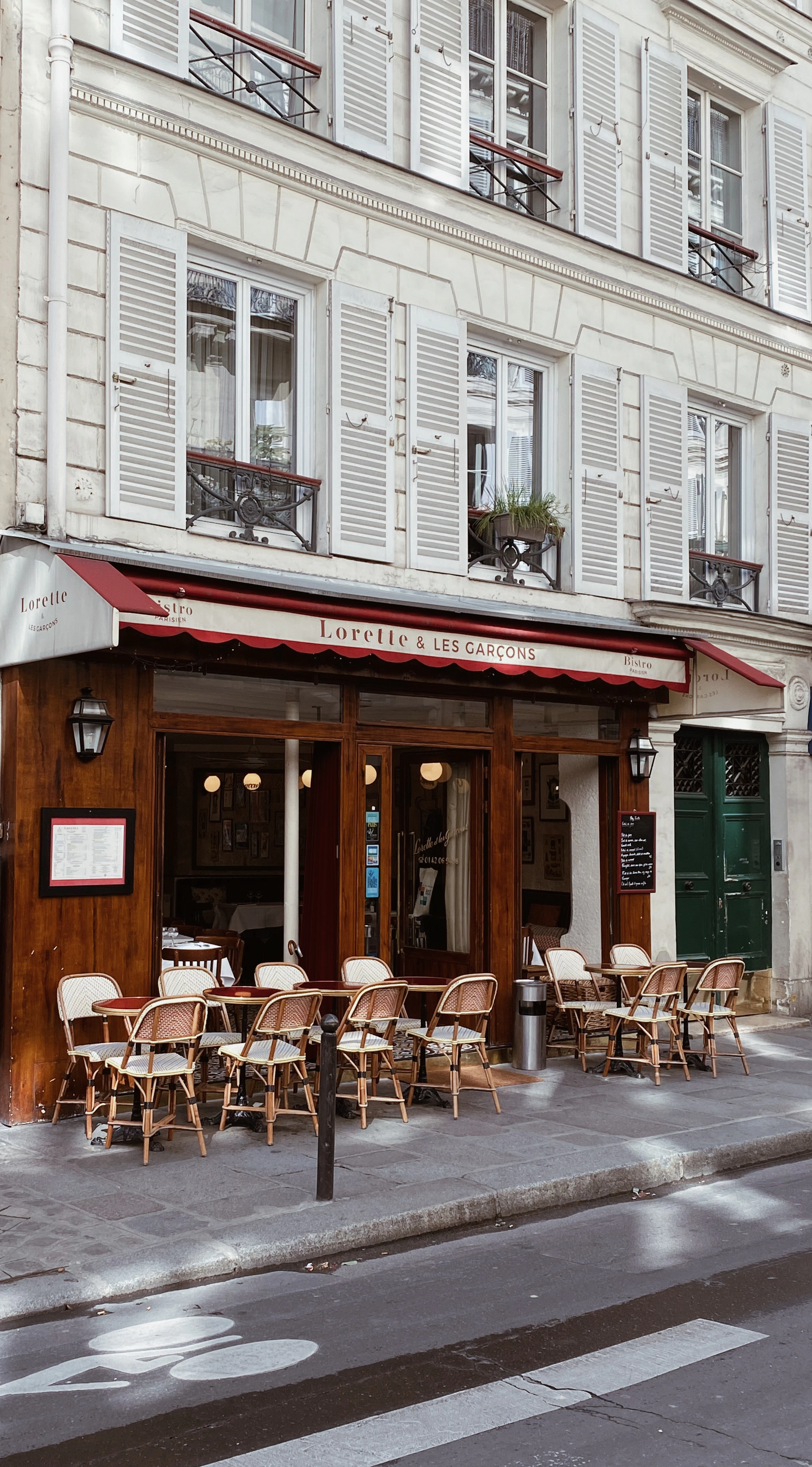 A Paris bistro terrace in the ninth arrondissement, its scalloped red awning lettered Lorette & les Garcons over woven rattan cafe chairs and small round tables on the pavement, below a white Haussmann facade of louvred shutters and iron balconies