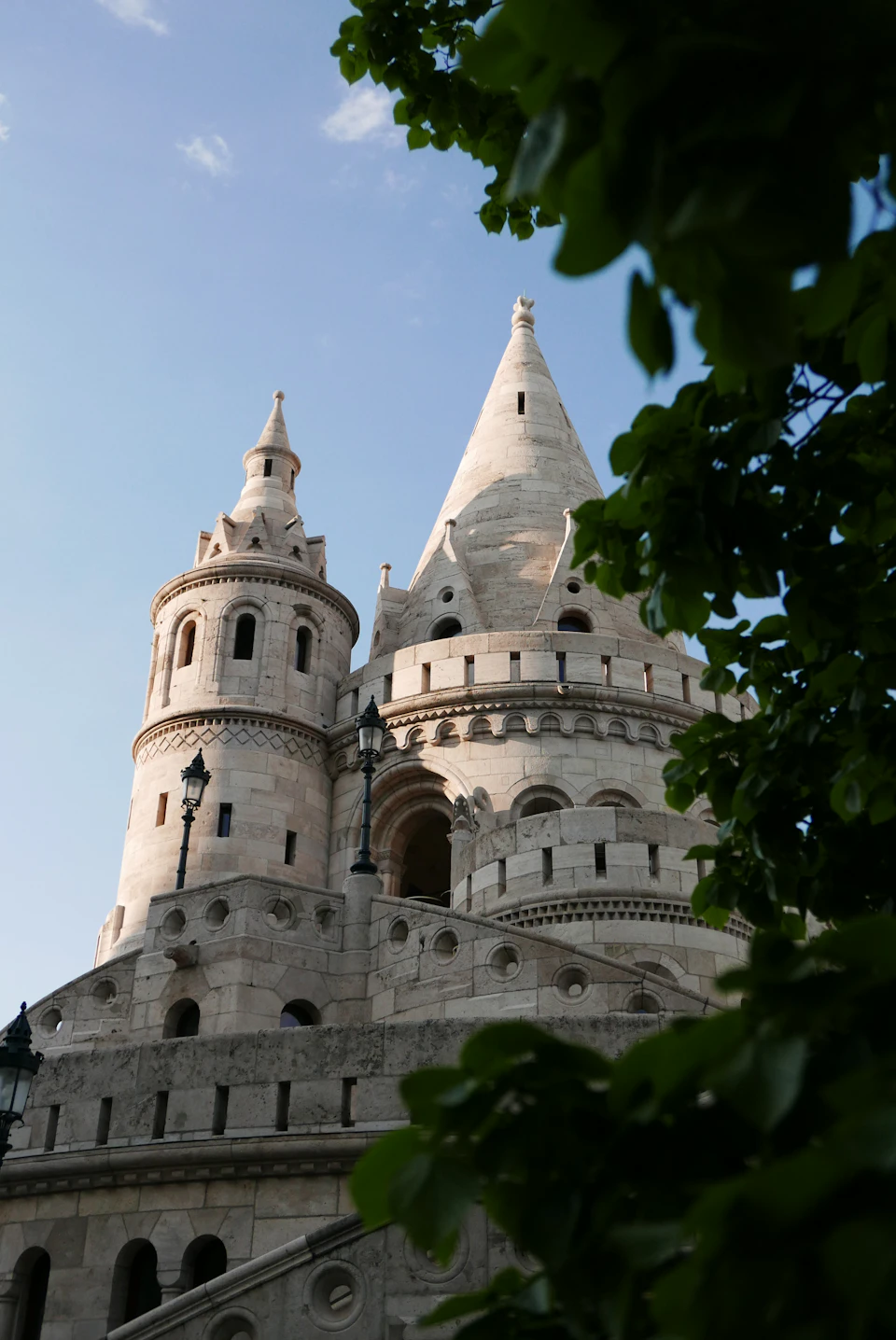 The image shows a majestic stone castle perched on a hill, with its multiple towers and spires reaching towards the sky. The castle is surrounded by lush greenery, including trees that frame the scene from below. A small building can be seen in the foreground of the image, adding depth to the overall composition.
