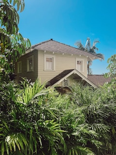 A two-story, beige house with a dark tiled roof is surrounded by lush tropical vegetation under a bright blue sky. The house has several windows with white trims, and palm trees are visible in the background.