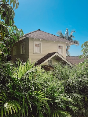A two-story, beige house with a dark tiled roof is surrounded by lush tropical vegetation under a bright blue sky. The house has several windows with white trims, and palm trees are visible in the background.