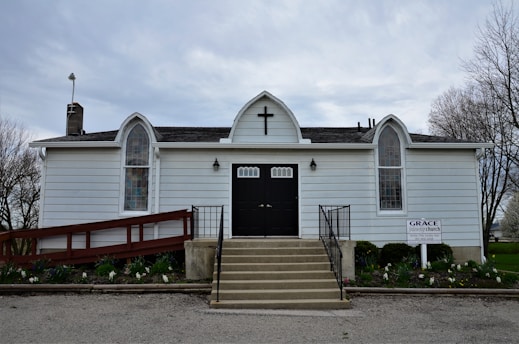A small white church building with a black door, positioned at the top of a short set of stairs. The building has a cross above the door and three arched windows with stained glass panels. There is a red ramp to the left side of the building and a sign that reads 'Grace Church' on the right. Surrounding the building is a modest garden with some flowers and a few trees without leaves visible in the background. The sky is overcast.