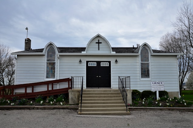 A small white church building with a black door, positioned at the top of a short set of stairs. The building has a cross above the door and three arched windows with stained glass panels. There is a red ramp to the left side of the building and a sign that reads 'Grace Church' on the right. Surrounding the building is a modest garden with some flowers and a few trees without leaves visible in the background. The sky is overcast.