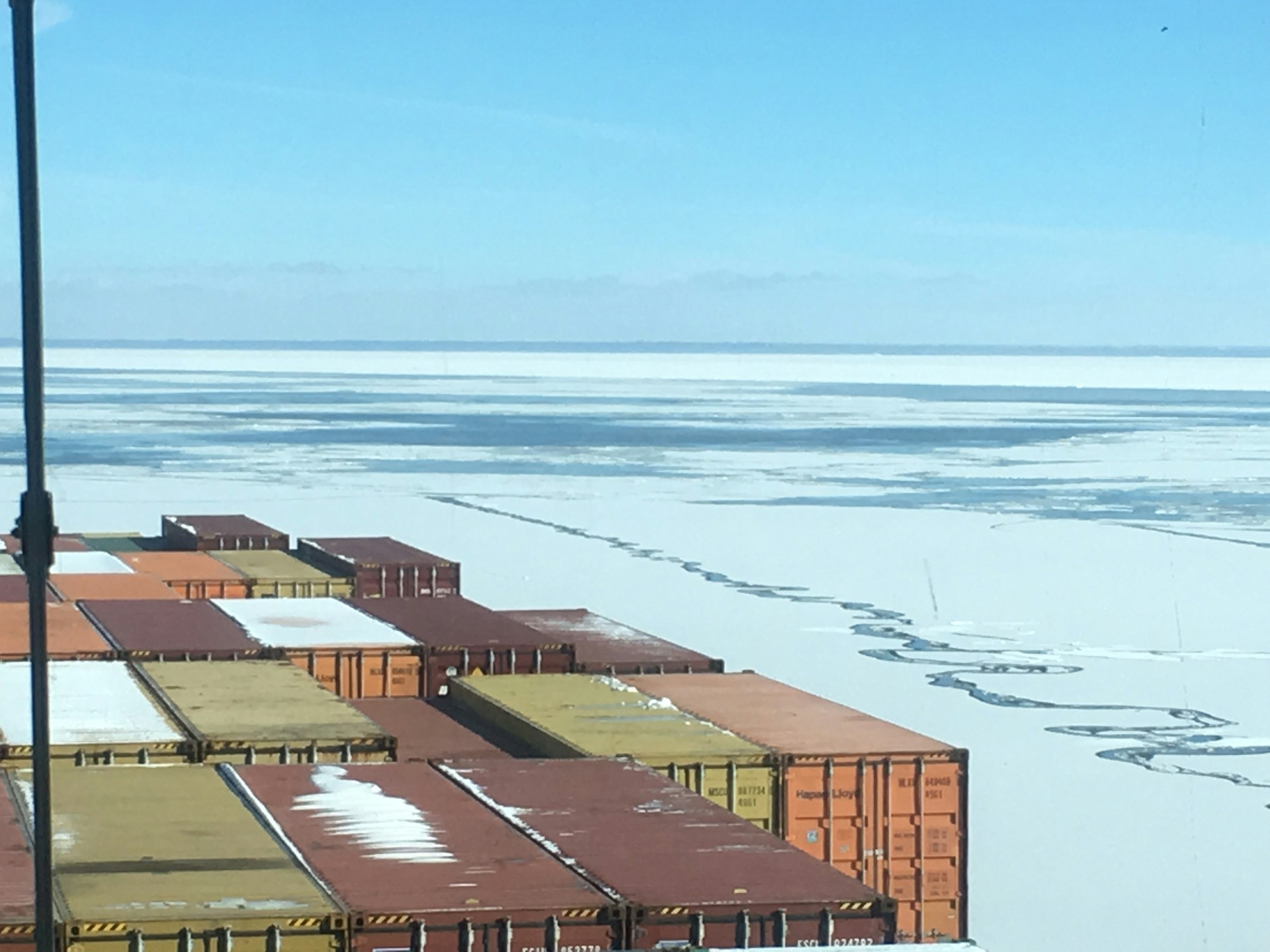 A view from a ship’s deck loaded with colorful shipping containers floating through icy waters. The horizon shows a vast expanse of frozen sea with patches of open water. The sky is clear with a light blue color.