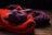 A bundle of yarn and knitted fabrics are laid out on a wooden surface. Specifically, a purple ball of yarn with two knitting needles inserted into it is placed on top of a red knitted fabric. Behind the red fabric, there is another piece of knitted material in a darker purple color.