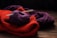 A bundle of yarn and knitted fabrics are laid out on a wooden surface. Specifically, a purple ball of yarn with two knitting needles inserted into it is placed on top of a red knitted fabric. Behind the red fabric, there is another piece of knitted material in a darker purple color.