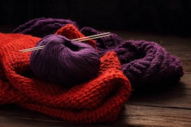 A bundle of yarn and knitted fabrics are laid out on a wooden surface. Specifically, a purple ball of yarn with two knitting needles inserted into it is placed on top of a red knitted fabric. Behind the red fabric, there is another piece of knitted material in a darker purple color.