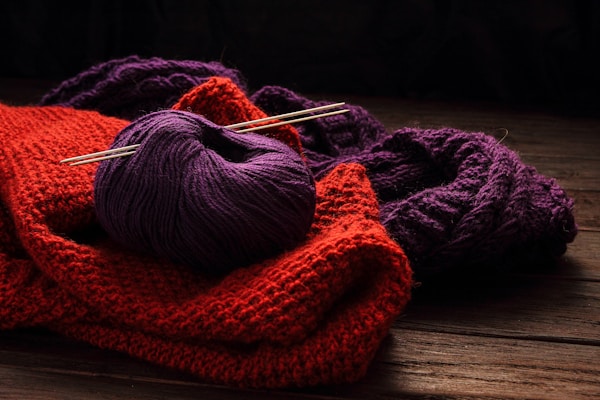 A bundle of yarn and knitted fabrics are laid out on a wooden surface. Specifically, a purple ball of yarn with two knitting needles inserted into it is placed on top of a red knitted fabric. Behind the red fabric, there is another piece of knitted material in a darker purple color.