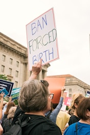 A group of people participating in a protest, holding signs and banners in front of large buildings. One prominent sign reads 'BAN FORCED BIRTH' in blue and green lettering. The crowd appears engaged and determined.