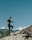 A person in black athletic wear is stepping onto a rocky ledge with mountains in the background. The sky is clear and blue, and there are some trees and shrubs around.