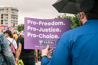 A group of people is gathered at a public demonstration holding signs. One prominent sign in the foreground is purple and reads 'Pro-Freedom. Pro-Justice. Pro-Choice.' The scene takes place outdoors with trees and a building visible in the background. The people are dressed in casual clothing and appear to be engaged in the protest.