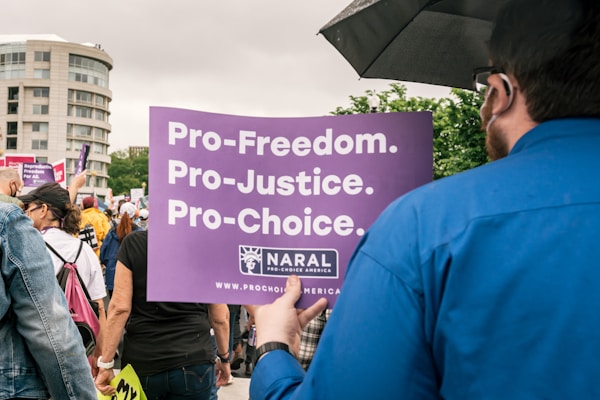 A group of people is gathered at a public demonstration holding signs. One prominent sign in the foreground is purple and reads 'Pro-Freedom. Pro-Justice. Pro-Choice.' The scene takes place outdoors with trees and a building visible in the background. The people are dressed in casual clothing and appear to be engaged in the protest.