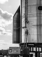 The image depicts a close-up view of an industrial structure made of metal, with various cables and components attached to it. In the foreground, there is portable equipment with an Atlas Copco label for rental services. The sky appears partly cloudy with a mix of light and darker clouds.