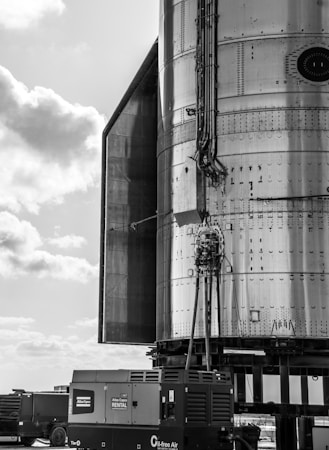 The image depicts a close-up view of an industrial structure made of metal, with various cables and components attached to it. In the foreground, there is portable equipment with an Atlas Copco label for rental services. The sky appears partly cloudy with a mix of light and darker clouds.