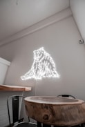 A room with a minimalist interior design features a small round wooden table and a chair. The wall glows with a neon light in the shape of a bear, creating a modern and artistic atmosphere. The walls are painted in a light neutral color, contributing to a clean and uncluttered look.
