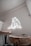A room with a minimalist interior design features a small round wooden table and a chair. The wall glows with a neon light in the shape of a bear, creating a modern and artistic atmosphere. The walls are painted in a light neutral color, contributing to a clean and uncluttered look.