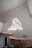A room with a minimalist interior design features a small round wooden table and a chair. The wall glows with a neon light in the shape of a bear, creating a modern and artistic atmosphere. The walls are painted in a light neutral color, contributing to a clean and uncluttered look.
