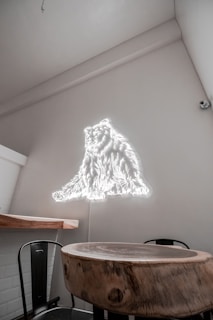 A room with a minimalist interior design features a small round wooden table and a chair. The wall glows with a neon light in the shape of a bear, creating a modern and artistic atmosphere. The walls are painted in a light neutral color, contributing to a clean and uncluttered look.