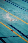A person is swimming in a clear blue swimming pool, performing a freestyle stroke. The pool is divided by yellow and blue lane markers, creating clear lanes for swimmer navigation. Water splashes around the swimmer, indicating motion and effort.
