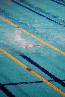 A person is swimming in a clear blue swimming pool, performing a freestyle stroke. The pool is divided by yellow and blue lane markers, creating clear lanes for swimmer navigation. Water splashes around the swimmer, indicating motion and effort.