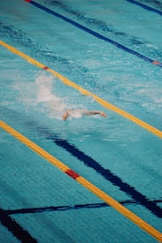 A person is swimming in a clear blue swimming pool, performing a freestyle stroke. The pool is divided by yellow and blue lane markers, creating clear lanes for swimmer navigation. Water splashes around the swimmer, indicating motion and effort.