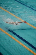 A swimmer is performing a butterfly stroke in an Olympic-sized swimming pool. The pool lanes are separated by floating lane markers, and the swimmer is wearing a white swim cap, goggles, and swimsuit. The water is a vibrant blue, with waves created by the swimming motion.