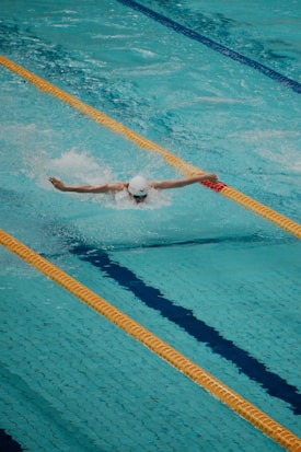 A swimmer is performing a butterfly stroke in an Olympic-sized swimming pool. The pool lanes are separated by floating lane markers, and the swimmer is wearing a white swim cap, goggles, and swimsuit. The water is a vibrant blue, with waves created by the swimming motion.