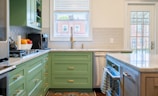 A modern kitchen interior features green cabinetry and a marble countertop. The backsplash is made of subway tiles, and there's a large window above the sink providing natural light. Appliances including a coffee maker and a gas stove are visible. A bowl of fresh fruit can be seen on the counter, and a tea towel hangs on the oven handle. There is a glass door leading to the outside.