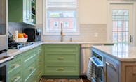 A modern kitchen interior features green cabinetry and a marble countertop. The backsplash is made of subway tiles, and there's a large window above the sink providing natural light. Appliances including a coffee maker and a gas stove are visible. A bowl of fresh fruit can be seen on the counter, and a tea towel hangs on the oven handle. There is a glass door leading to the outside.
