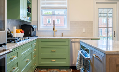 A modern kitchen interior features green cabinetry and a marble countertop. The backsplash is made of subway tiles, and there's a large window above the sink providing natural light. Appliances including a coffee maker and a gas stove are visible. A bowl of fresh fruit can be seen on the counter, and a tea towel hangs on the oven handle. There is a glass door leading to the outside.
