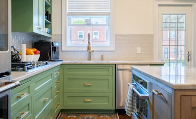A modern kitchen interior features green cabinetry and a marble countertop. The backsplash is made of subway tiles, and there's a large window above the sink providing natural light. Appliances including a coffee maker and a gas stove are visible. A bowl of fresh fruit can be seen on the counter, and a tea towel hangs on the oven handle. There is a glass door leading to the outside.